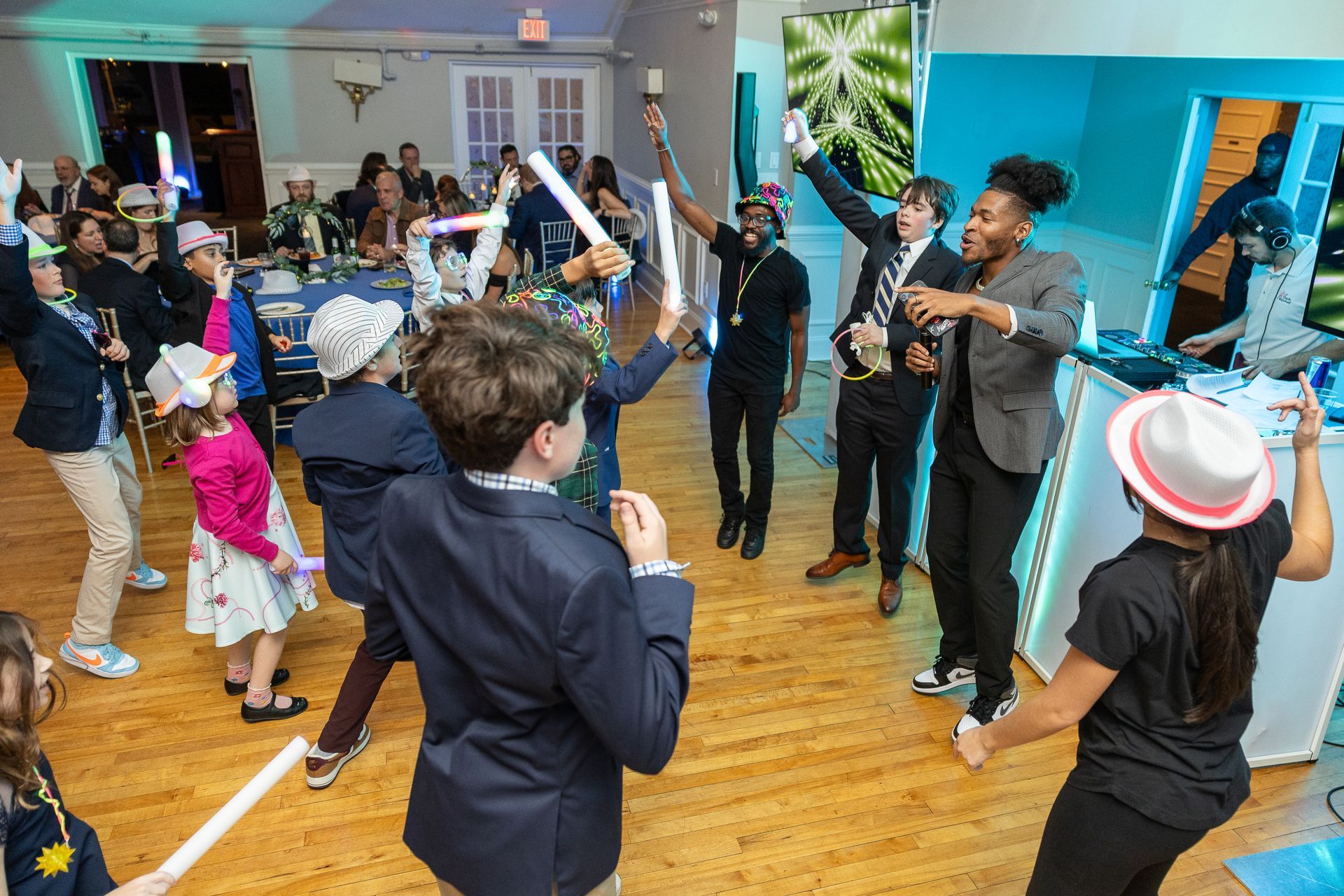 People dance and hold light-up foam sticks at an indoor party with tables, a DJ booth, and wooden flooring.