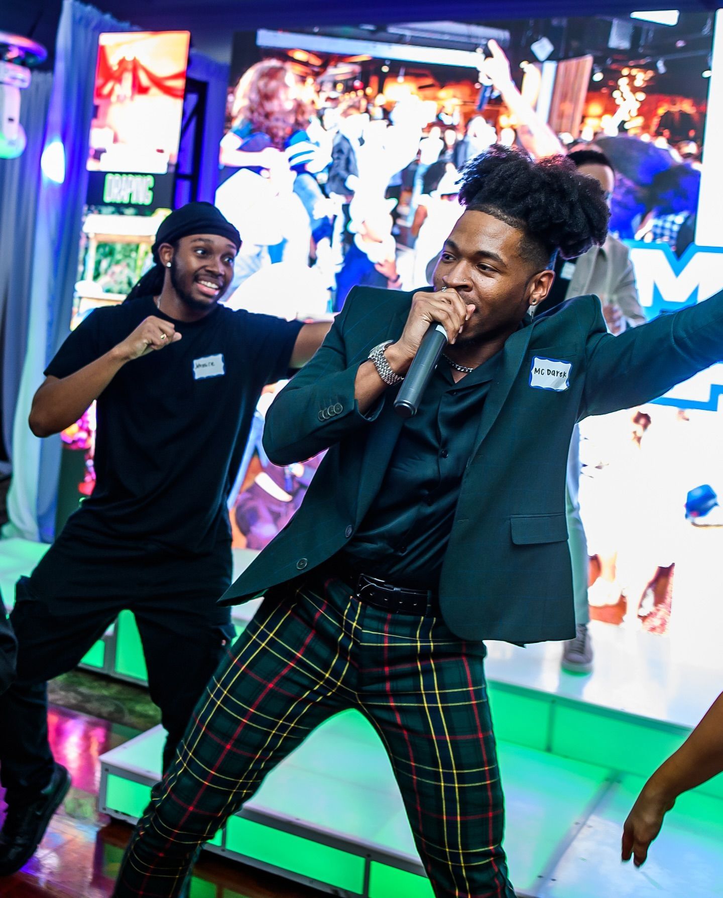 A person in a green blazer and plaid pants performs with a microphone.