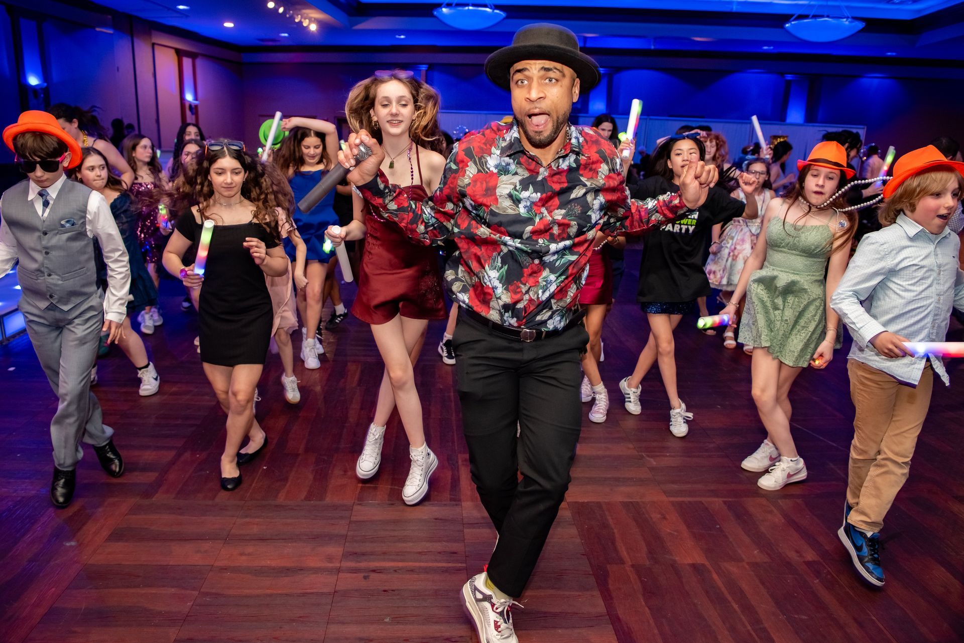 A person performs a backflip over a person kneeling in a blue dress at a party with guests watching.