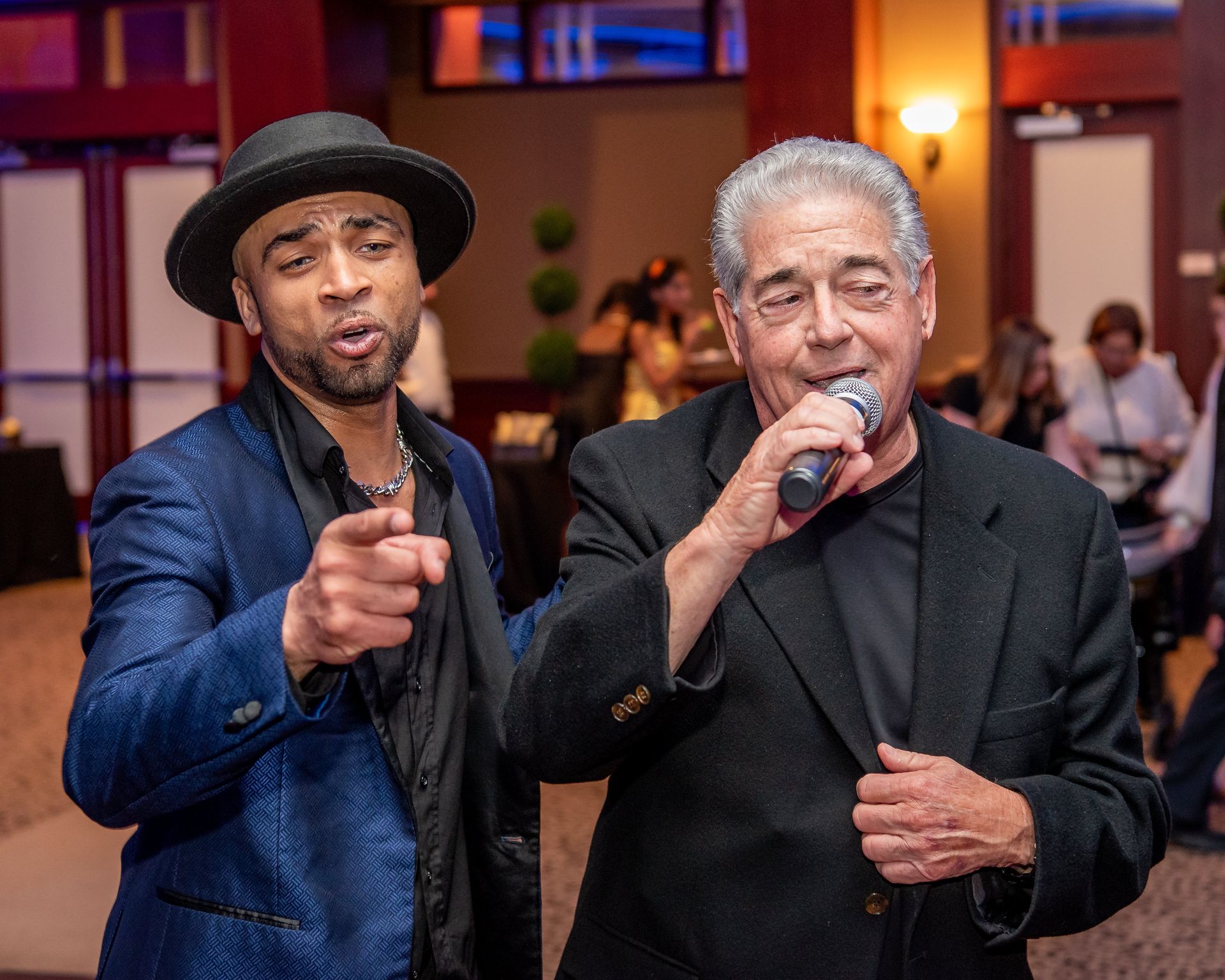 A man in a blue blazer and hat points forward while standing next to another man singing into a microphone at an event.