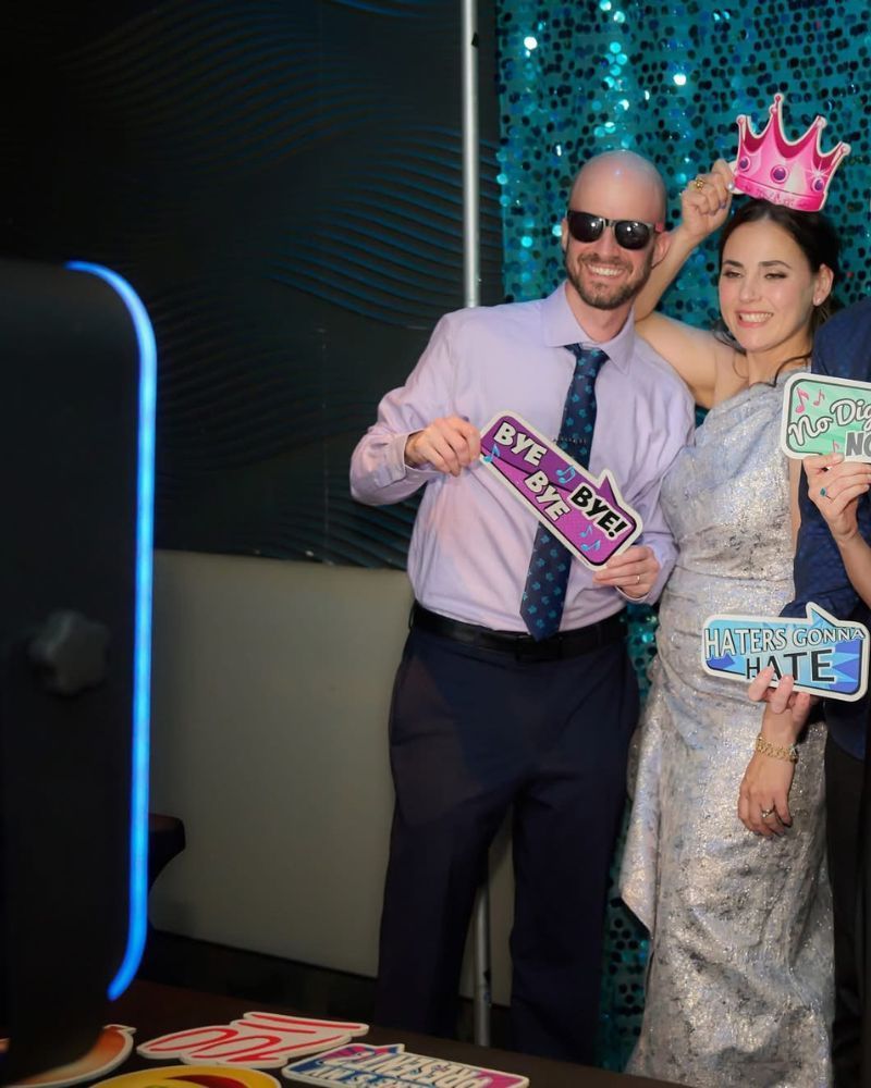 Couple posing with fun props in an open-air photo booth, smiling guests enjoying the lively atmosphere and vibrant backdrop at an event.