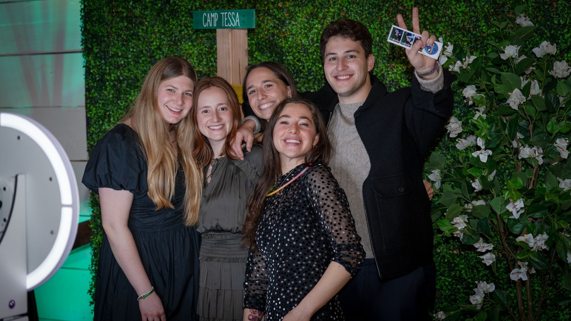 Group of guests posing in front of a green hedge backdrop using a photo booth with printed photo strips at a themed event.