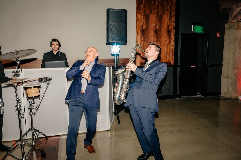 Man sings while saxophone player performs on dance floor with DJ in background.