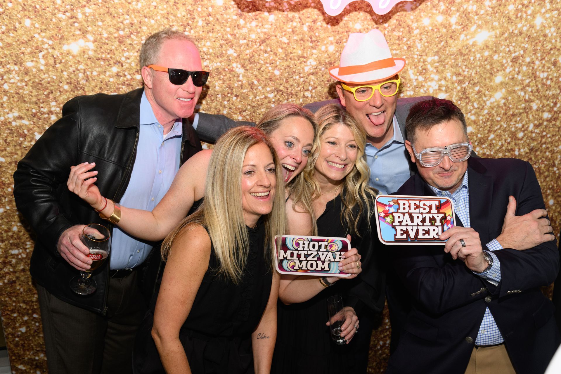 Adults posing with fun props and sunglasses in front of a gold glitter photo booth backdrop at a Mitzvah celebration.