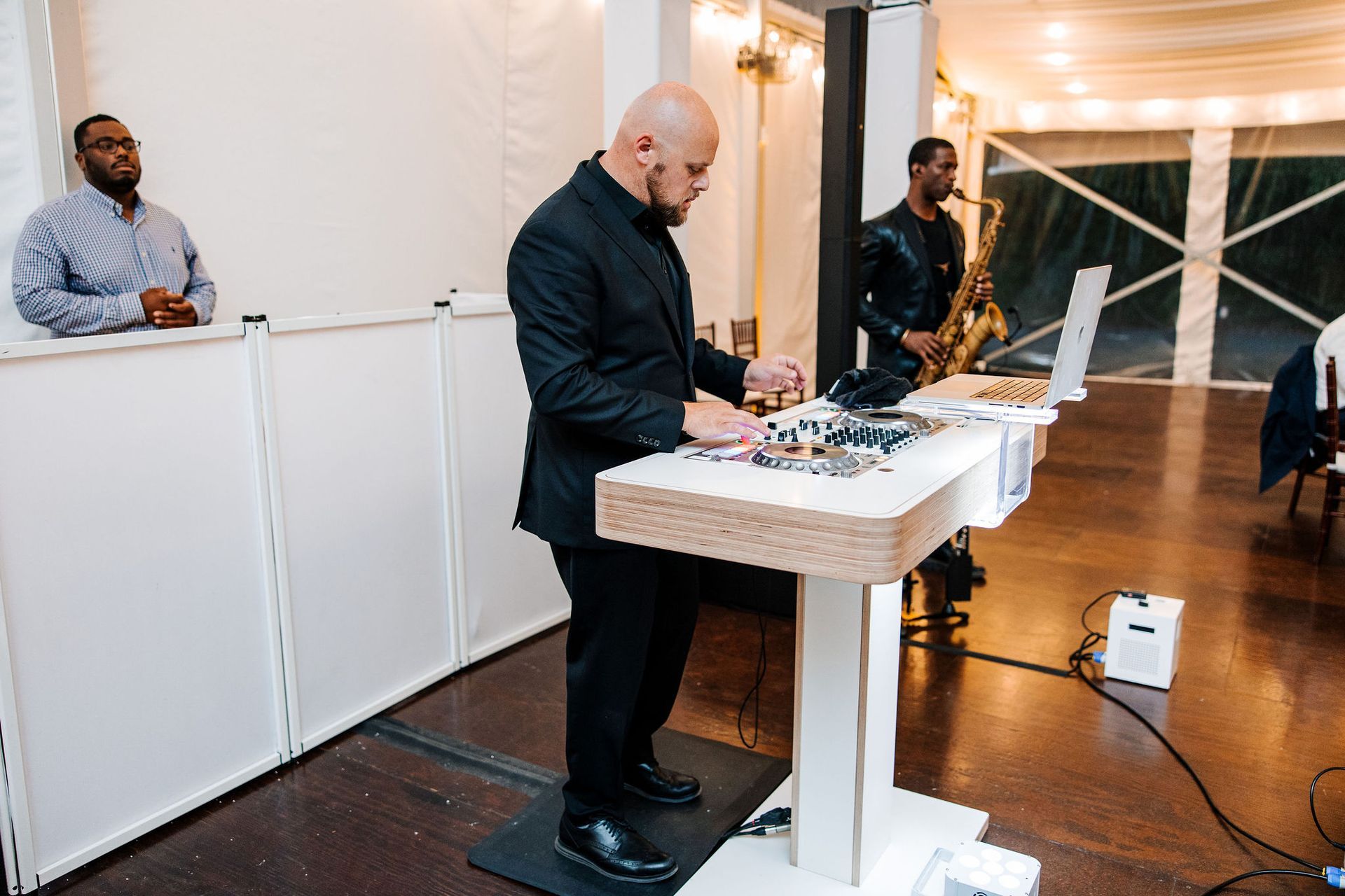 DJ in black suit at event with musician playing saxophone.