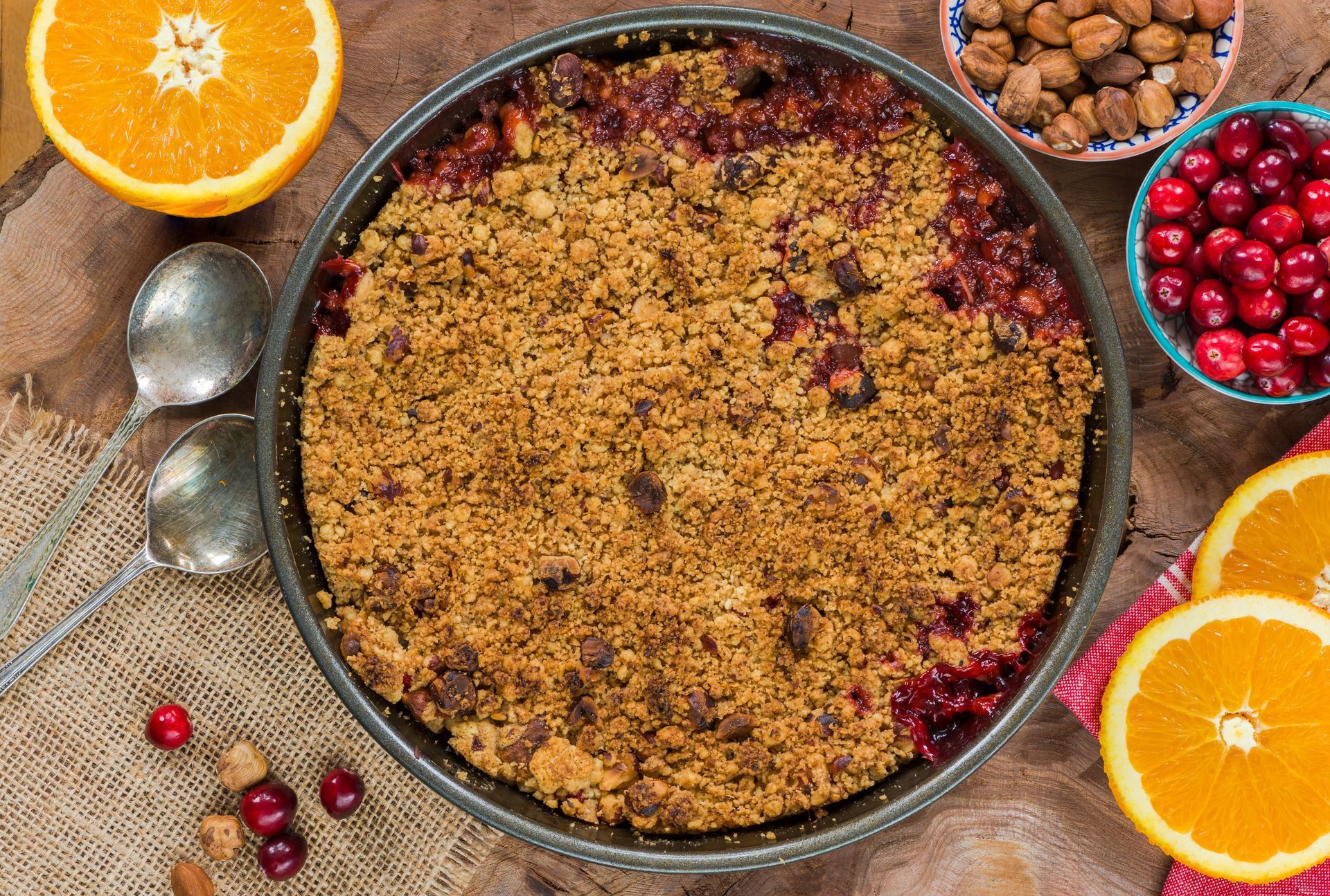 Cranberry crumble dessert in a round pan, surrounded by oranges, cranberries, and nuts.