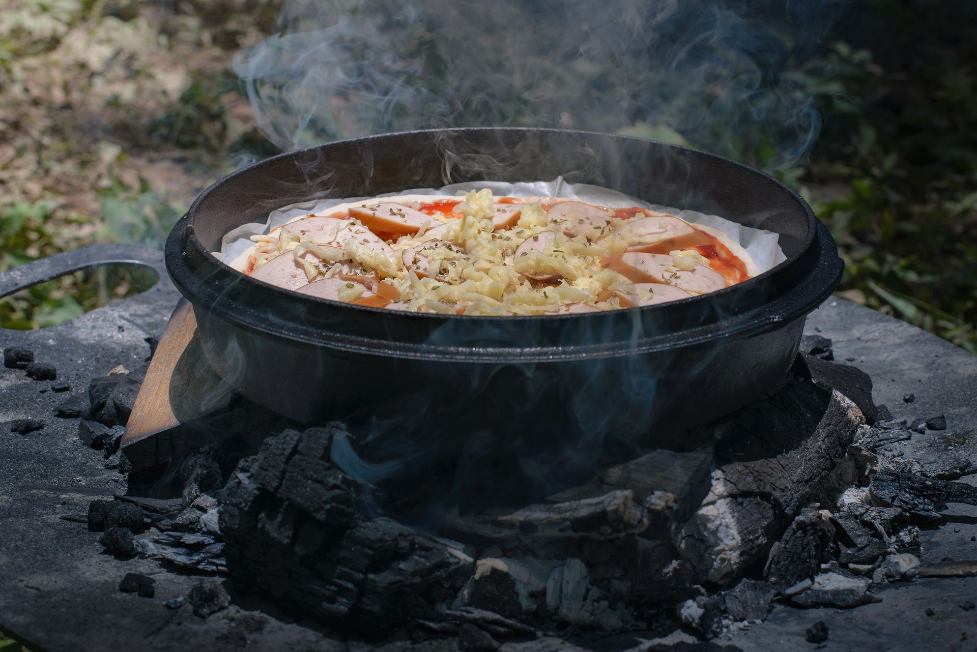 Pizza cooking in a black Dutch oven over charcoal outdoors, emitting steam.