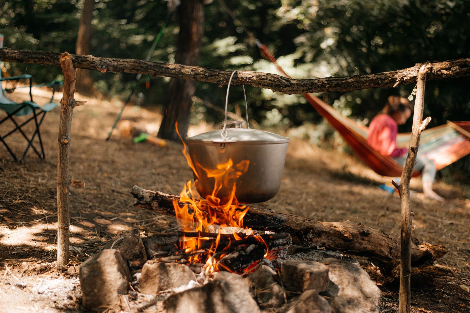 Campfire with pot hanging above flames, person in hammock in background.