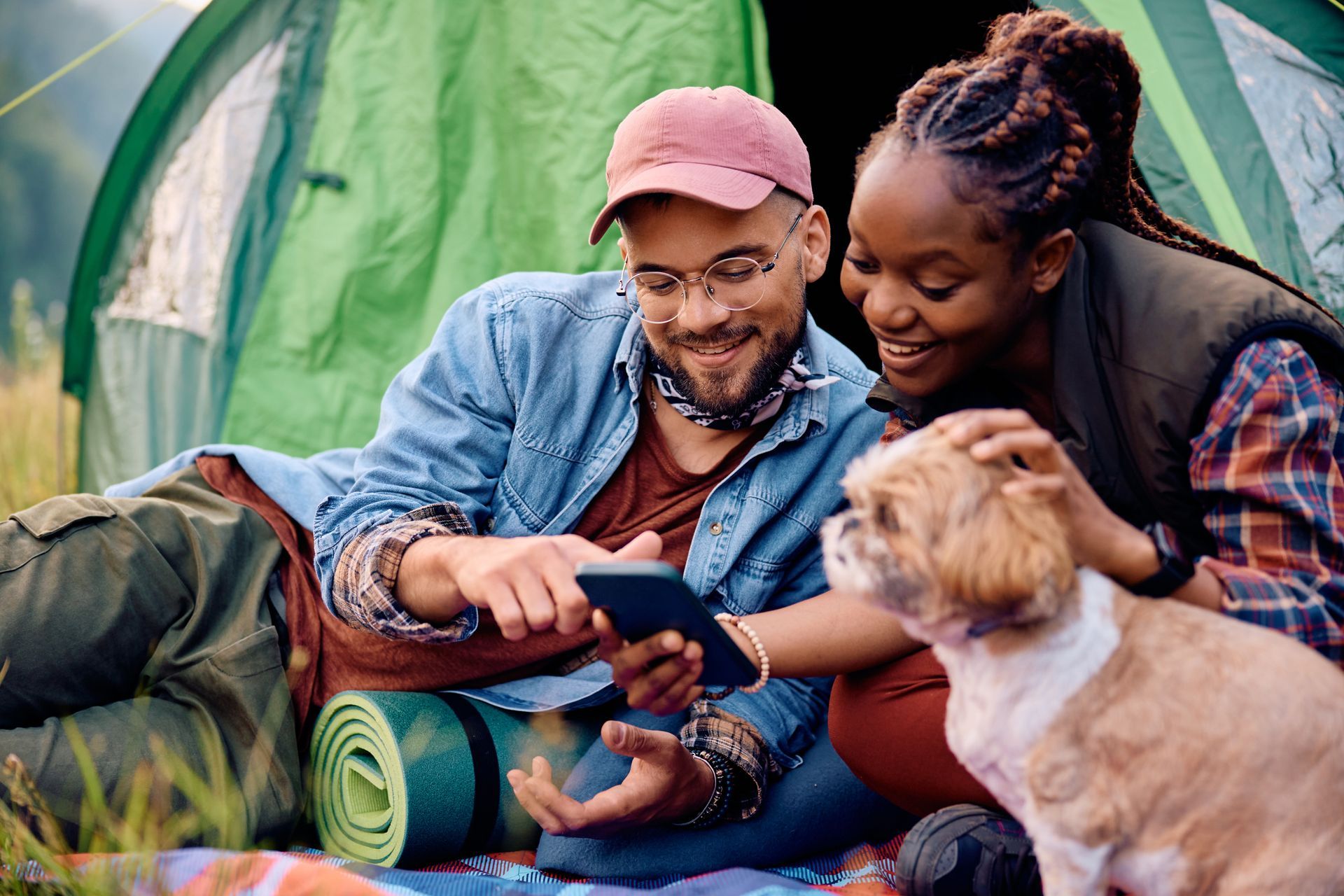 A man and a woman are laying in front of a tent with a dog and looking at a tablet.