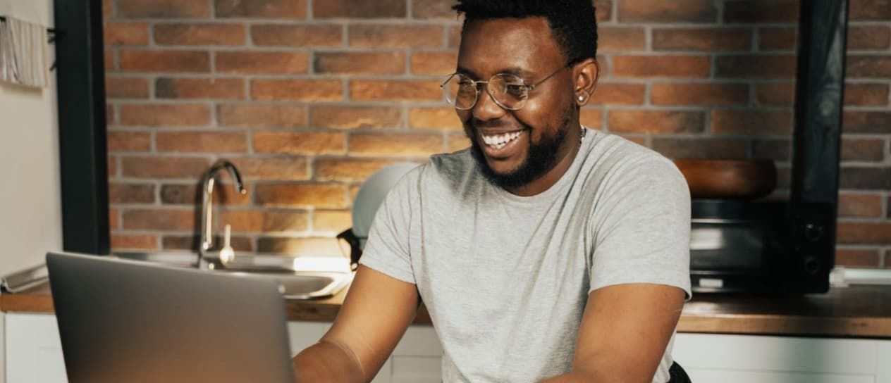 Man with glasses smiles while looking at laptop in a kitchen.