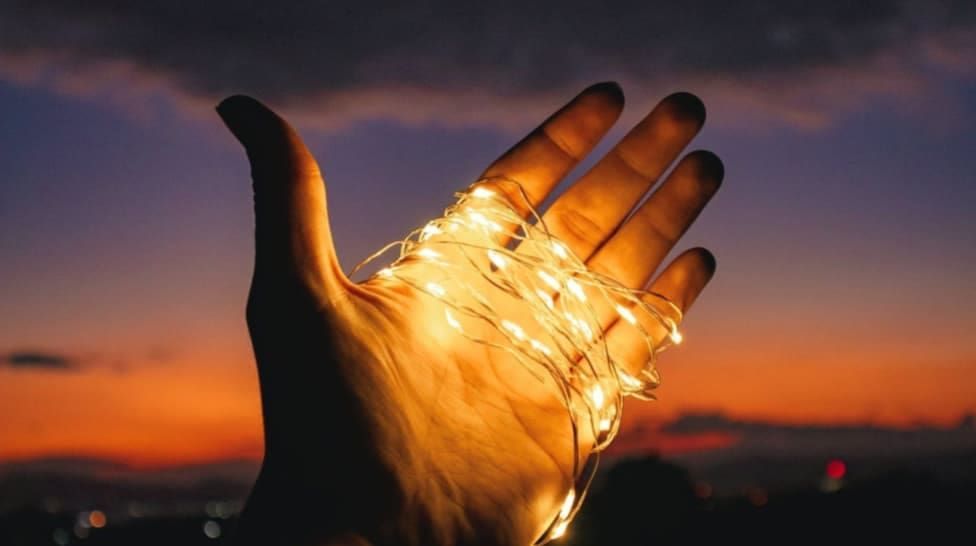 Hand holding glowing string lights against a sunset.