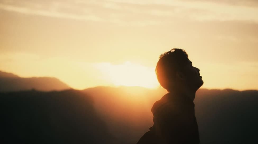 Person silhouetted against a sunset, gazing upward, with mountains in the background.