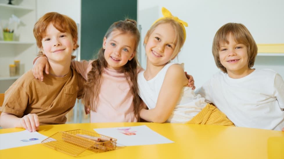 Four smiling children sit at a yellow table with art supplies, posing together.