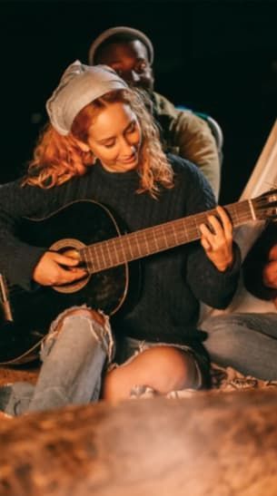 Woman with curly red hair plays a guitar around a campfire with friends at night.