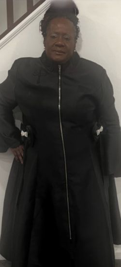 Woman in black coat, standing indoors with hand on hip.