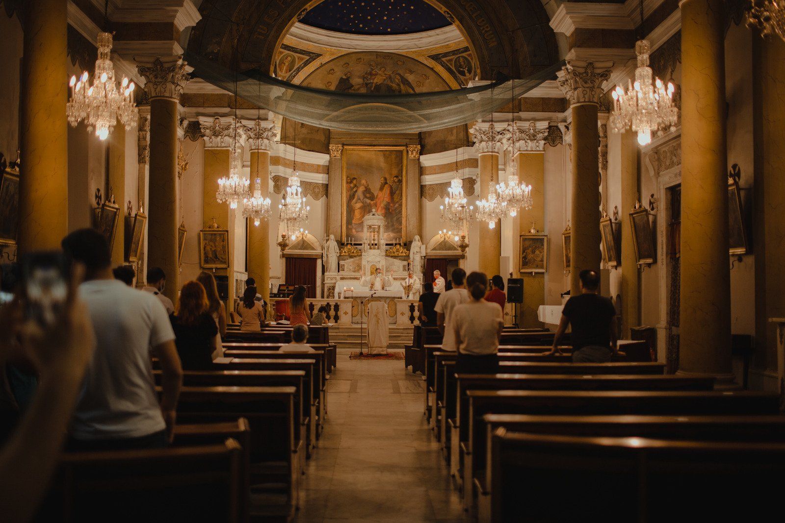 People in a church during a ceremony. Rows of pews face the altar, lit by chandeliers and ceiling lights.