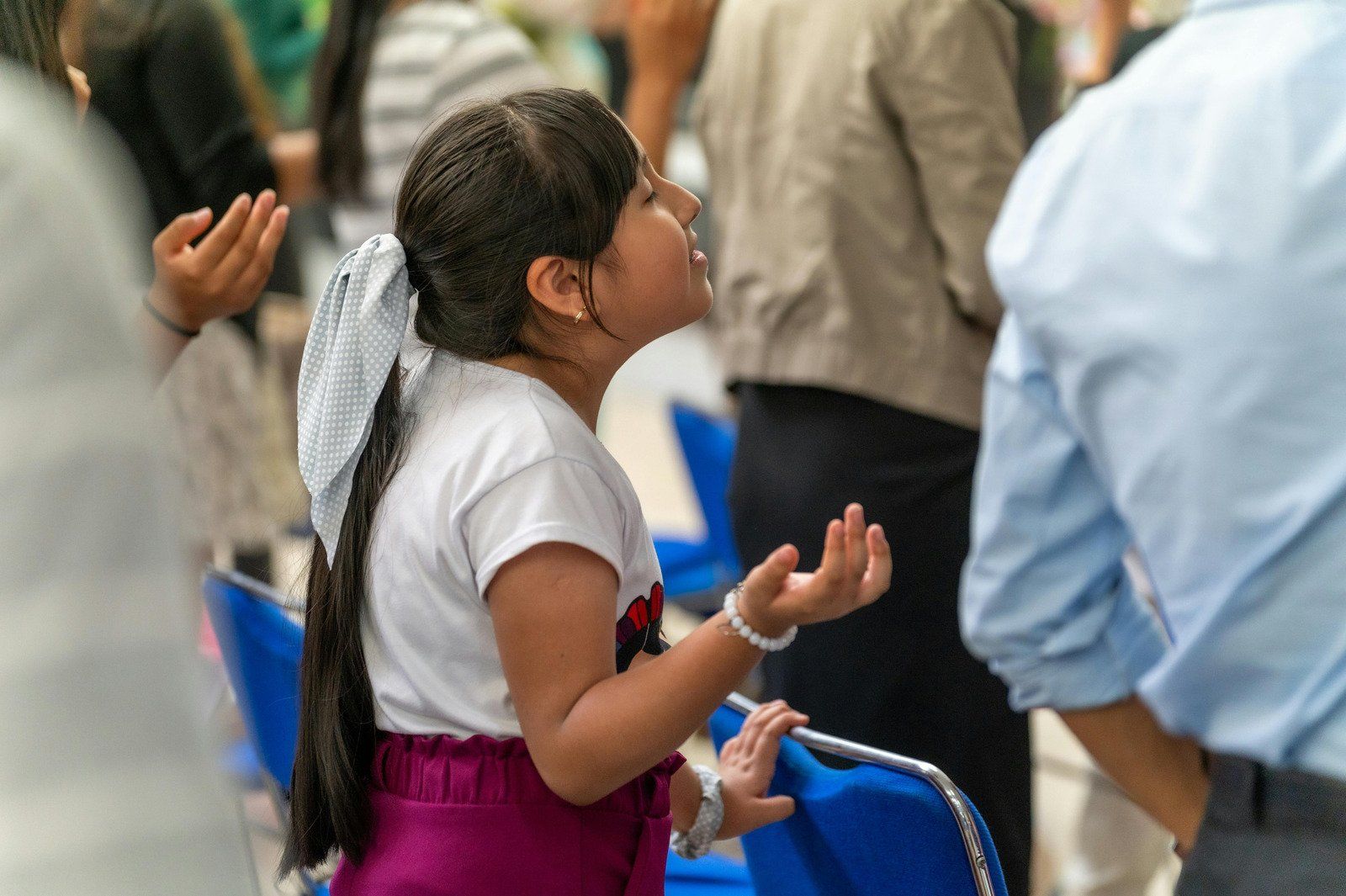 Girl praying with hands raised, wearing white shirt and purple skirt. People standing in background.