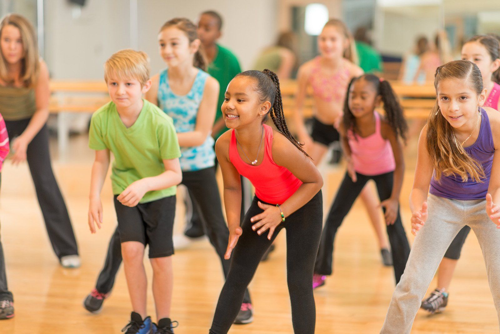 Children in colorful clothes dancing in a studio with a woman in the background.