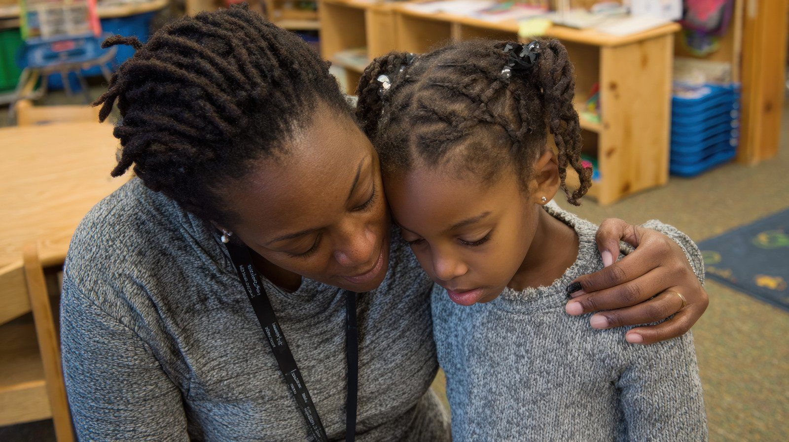 Woman comforts a young girl in a classroom; both have dark skin and are wearing gray sweaters.