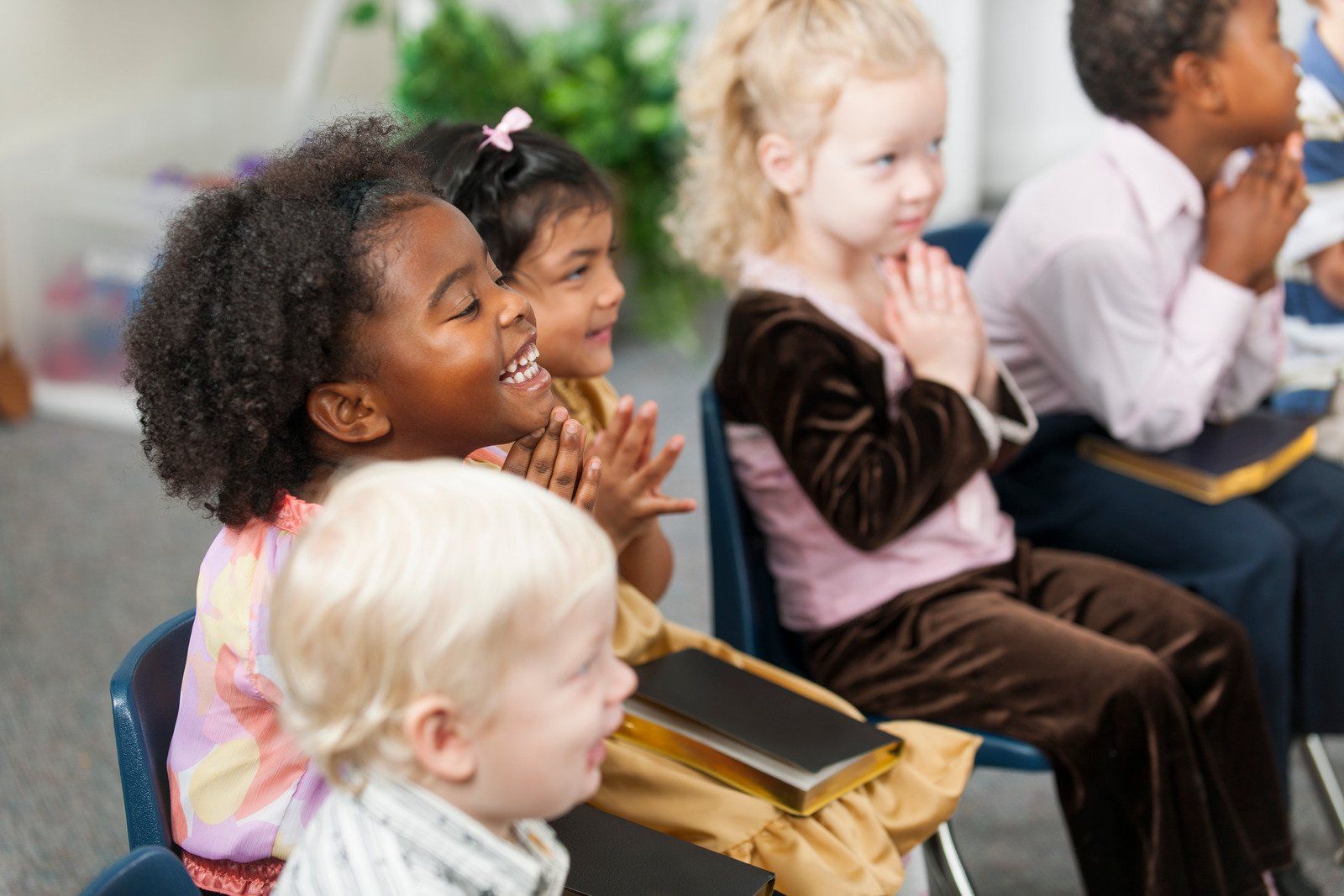 Children in a church, clapping and smiling, books on laps. Diverse group, light-filled room, soft colors.