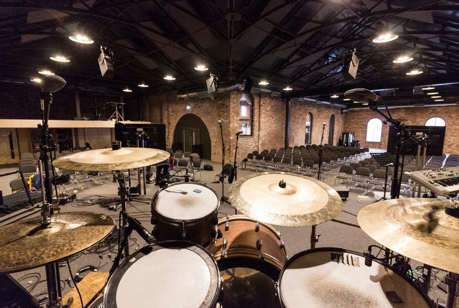 Drum set in a large hall with brick walls, stage, and rows of chairs, ready for a performance.
