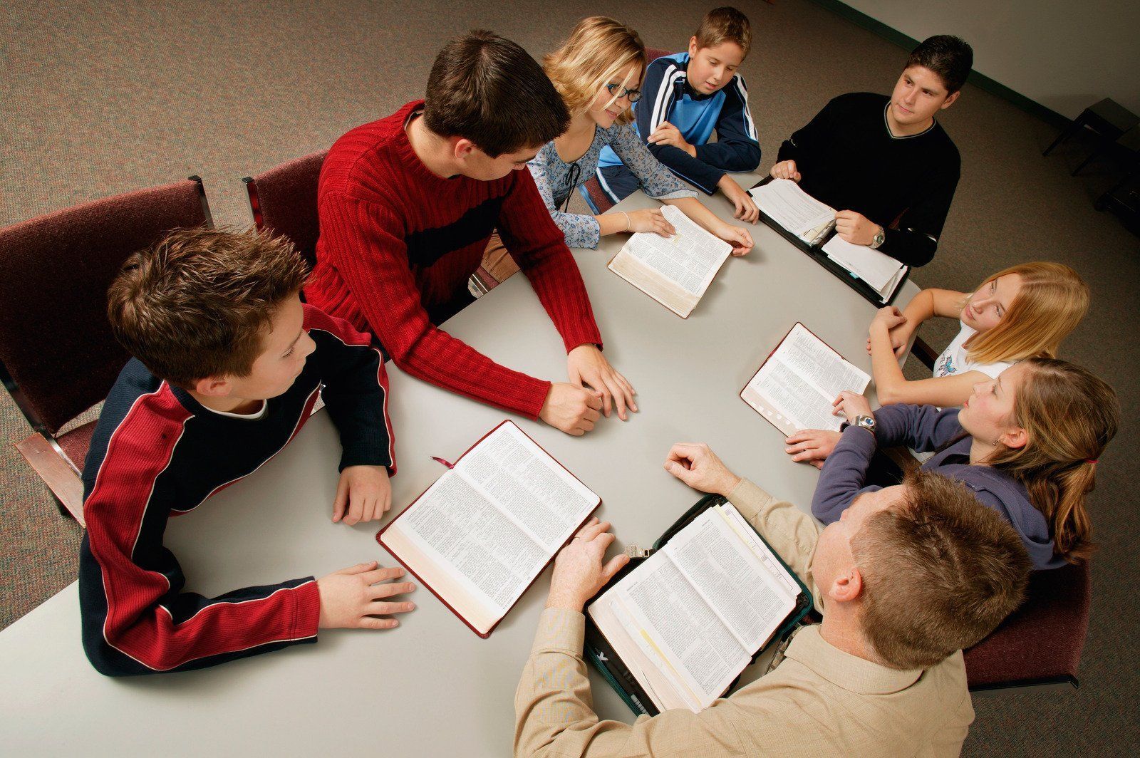 Group of young people around a table, studying open books, likely a bible study.