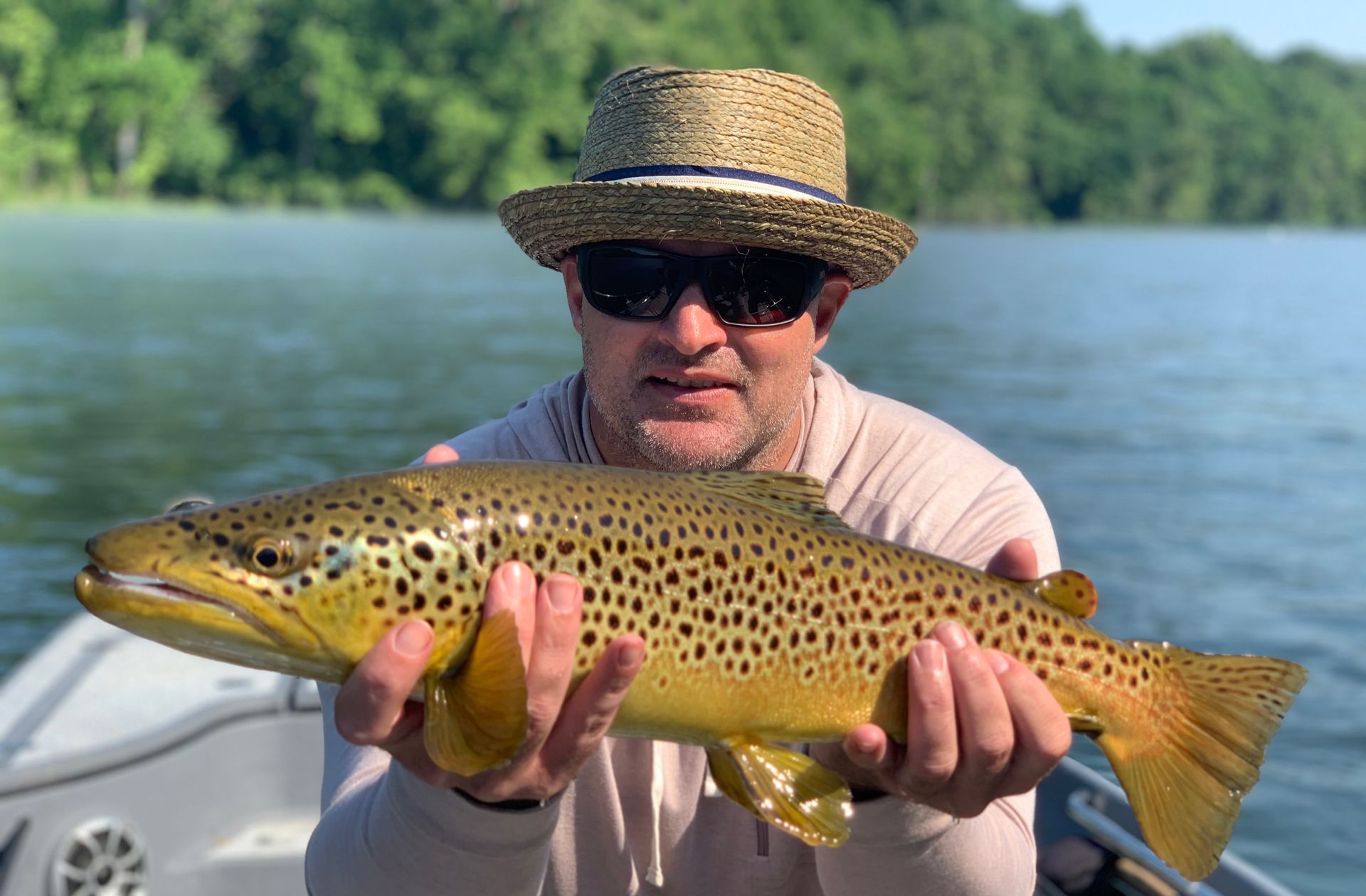 A man in a straw hat is holding a brown trout in his hands.