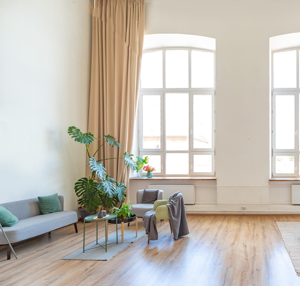 Timber floored lounge with minimalist couch, chairs and coffee tables on small rug. Green plants and green cushions backed by floor to ceiling drapes pulled aside to reveal large windows with light flowing through.