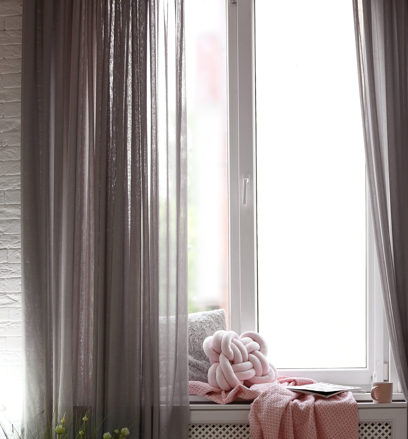 Close up of window with soft cushion and rug on sill, backed by purple toned semi-opaque sheer curtains.