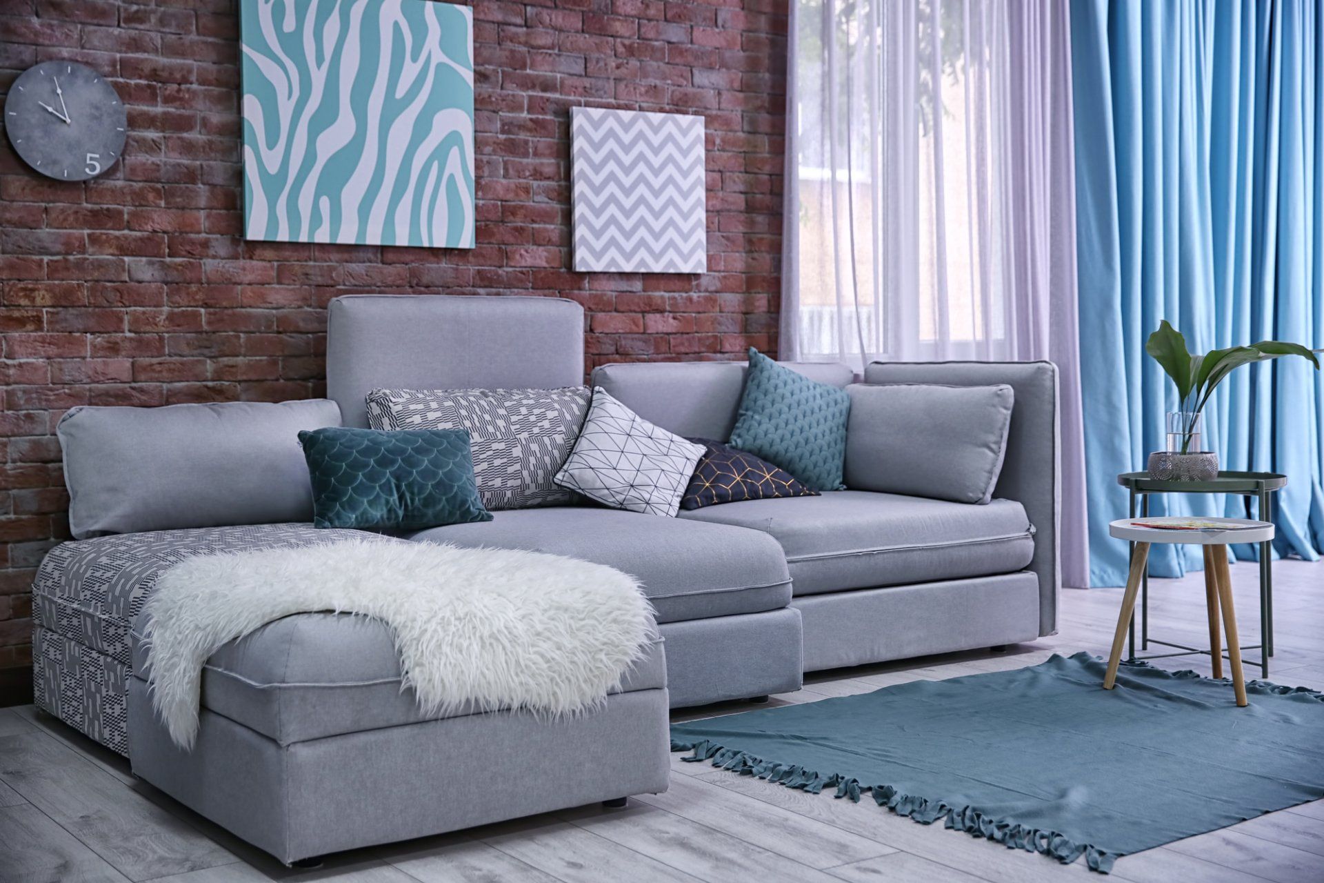 Brick walled lounge with grey couch covered with multiple cushions. Aqua toned art on wall complementing aqua run and drapes pulled aside to show soft sheers behind.