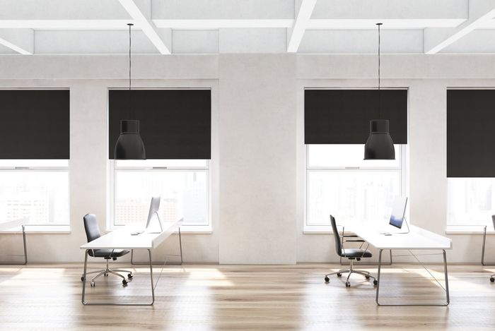 Ultra modern minimalistic white toned office setting showing 2 desks computers backed by 4 windows with black roller blinds pulled half way down.