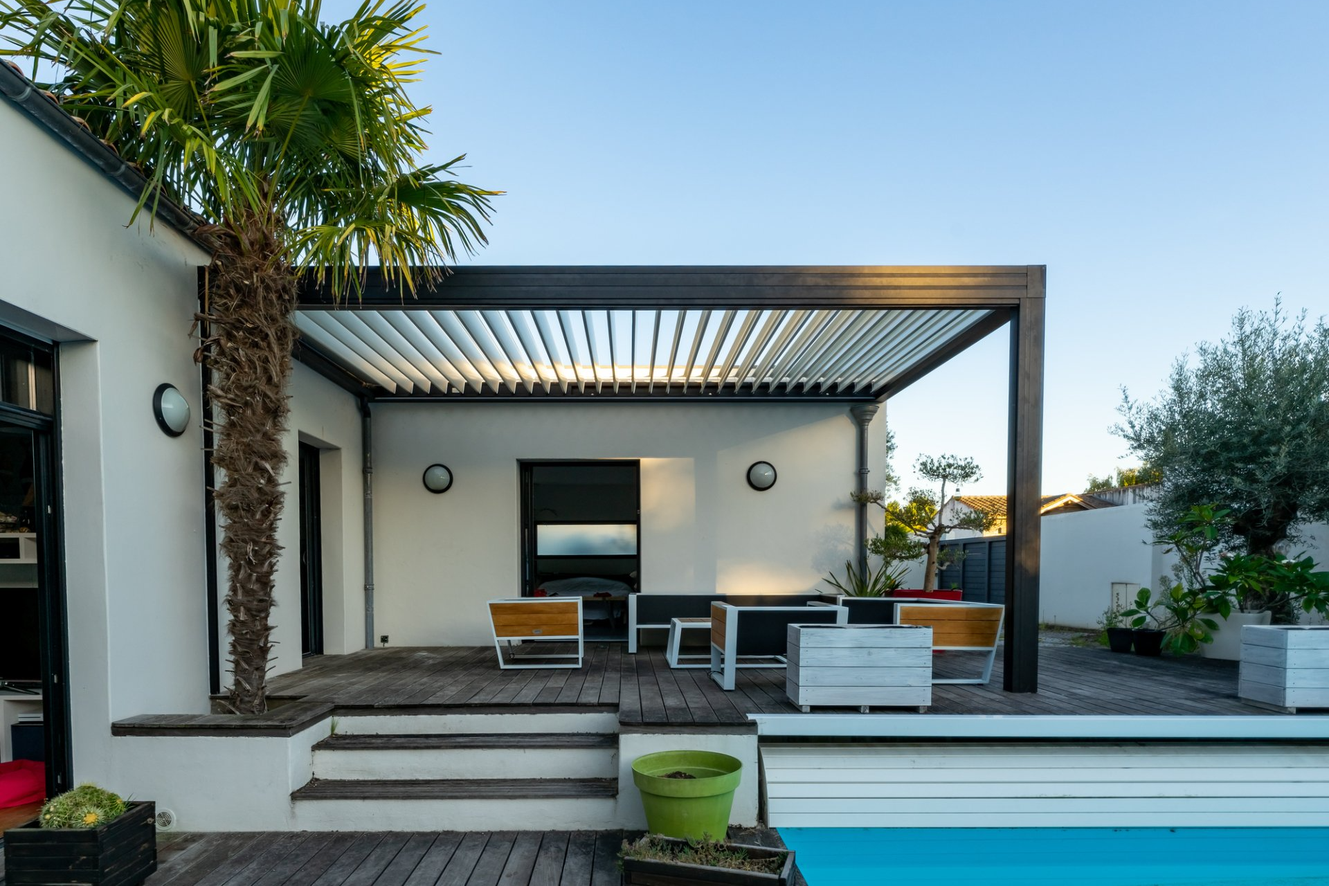 Fixed metal awning in place over outdoor entertaining area with timber and metal chairs, in front of swimming pool. Framed by tall palm tree.