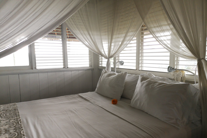 Close up of bed in beach house style bedroom surrounded by tied back opaque curtains exposing open plantation shutters.