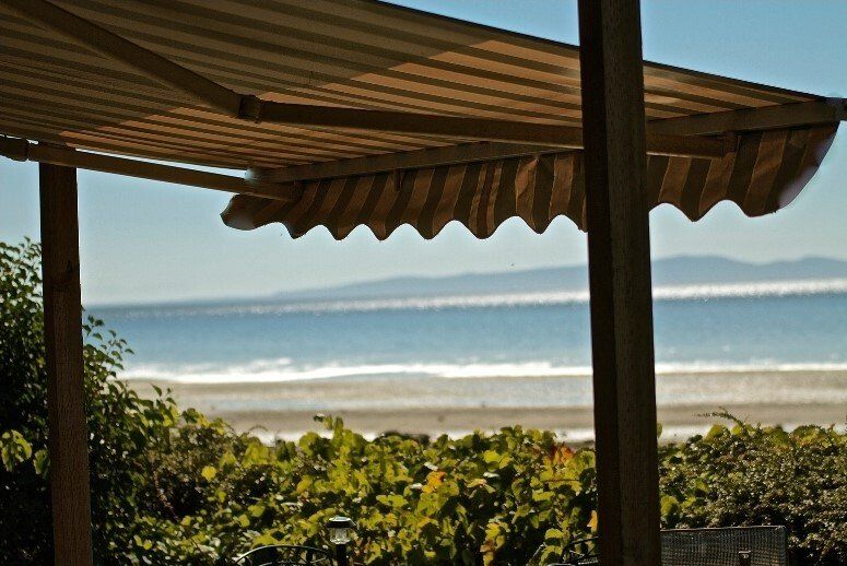 Outdoor awning extending out over hedge, with beach and distant mountains in the background.