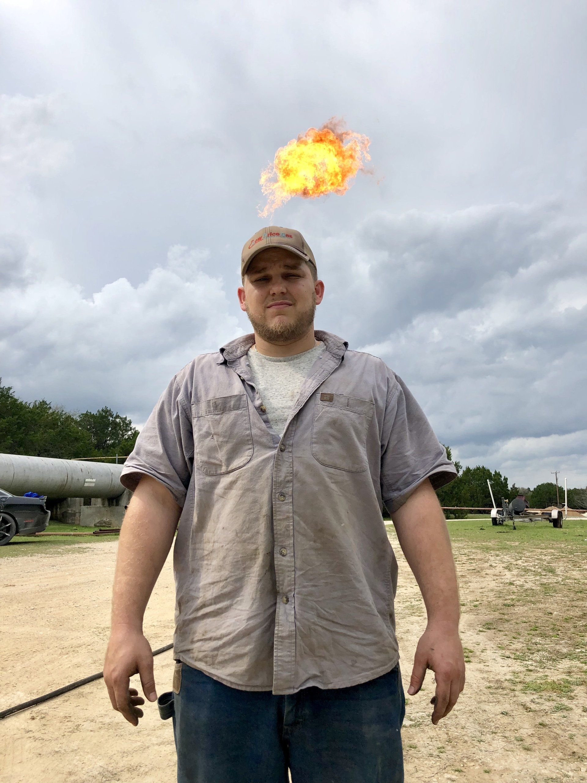Man with fire erupting from his head, wearing work clothes, standing outside on a cloudy day.