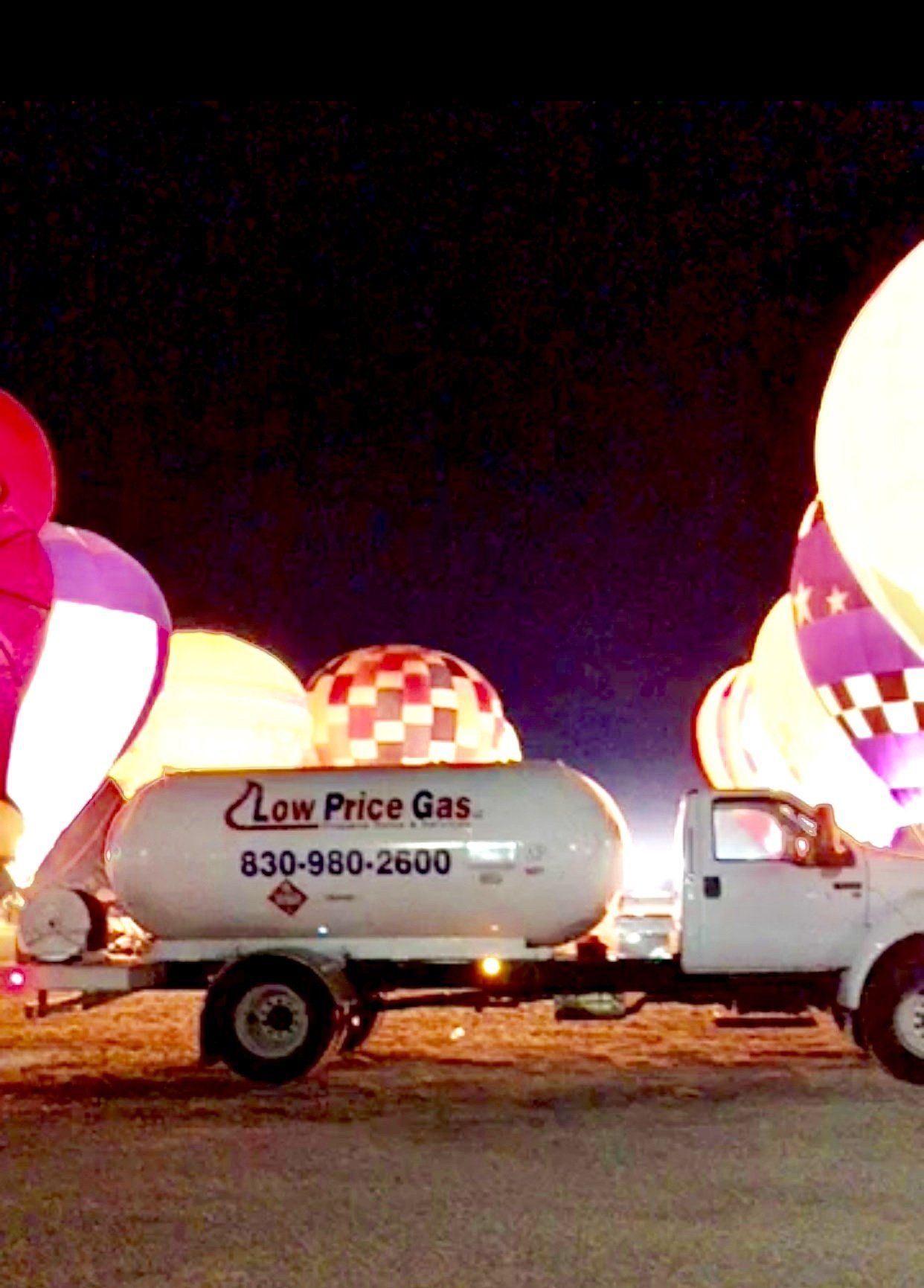 White truck with a gas tank in front of lit up hot air balloons at night. The tank has 