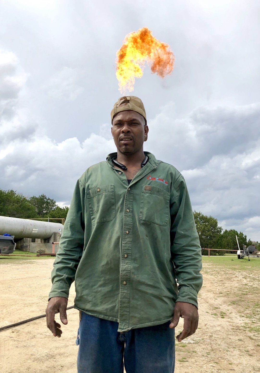 Black man with flames erupting from head, standing outdoors, green shirt, cloudy sky.