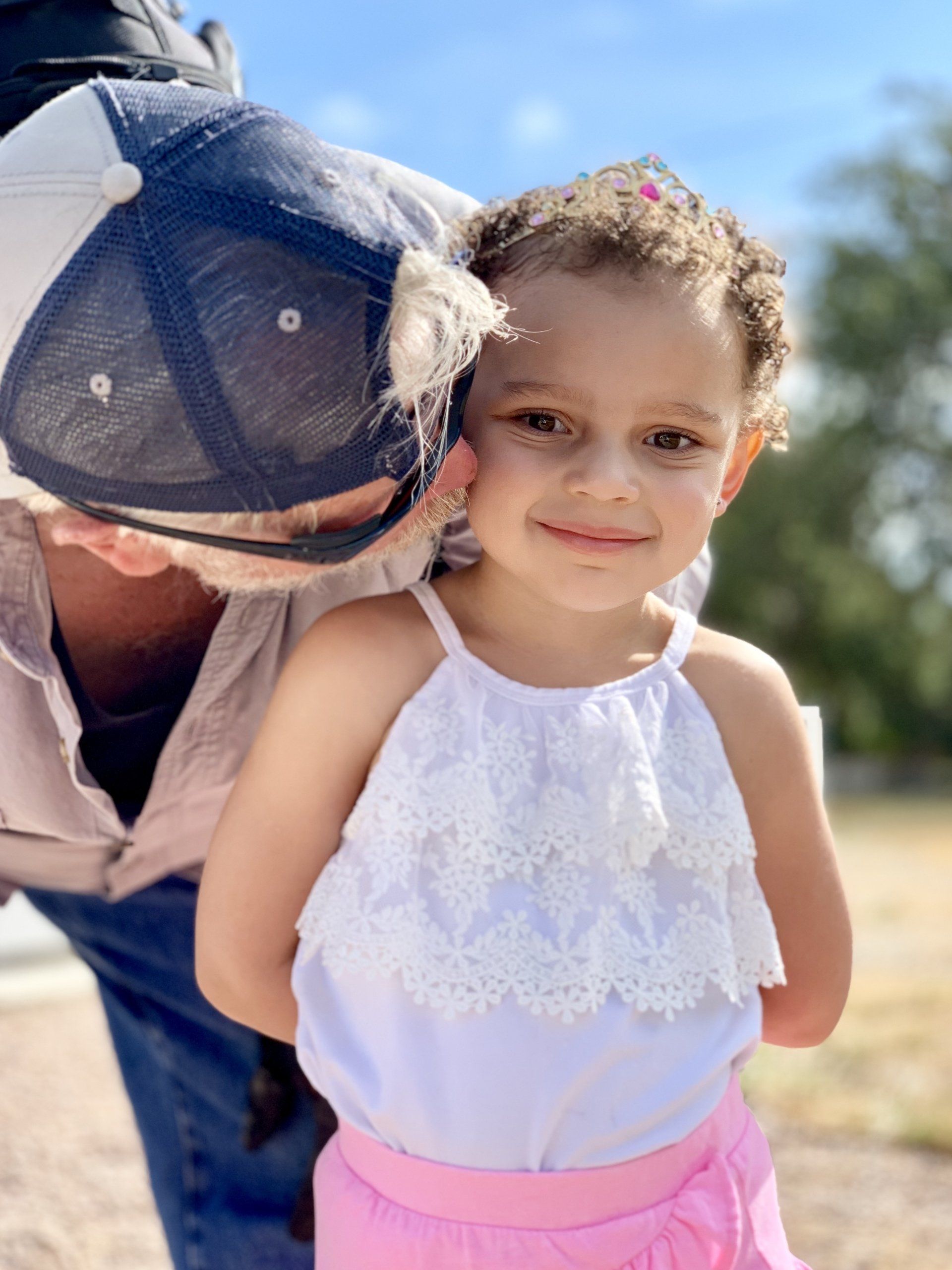 Young girl in pink skirt and white top smiles at the camera with a person in a hat partially in the frame.