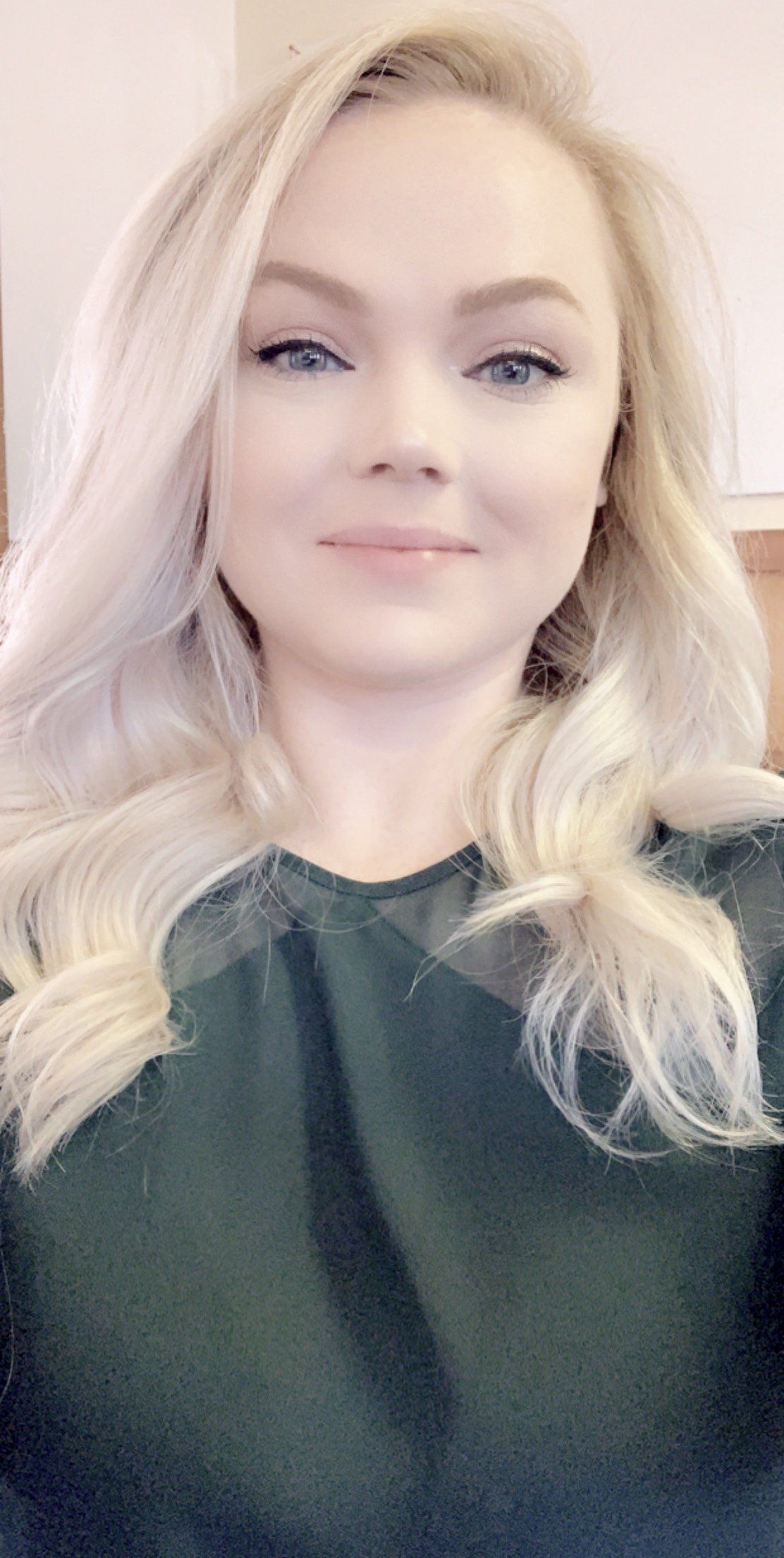 Blond woman with blue eyes smiles. She wears a dark green top.