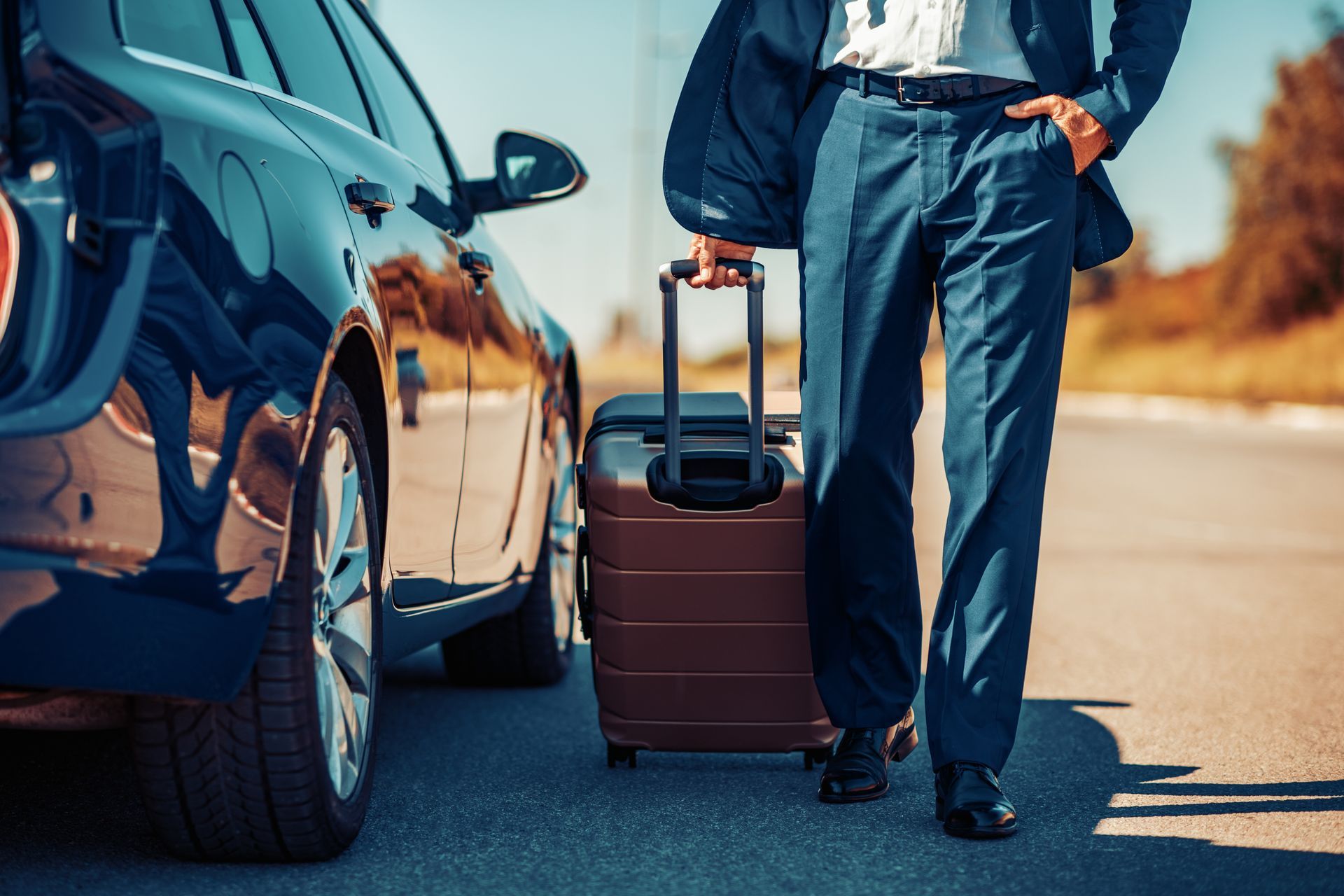 Man in suit with a suitcase next to a black car on a road