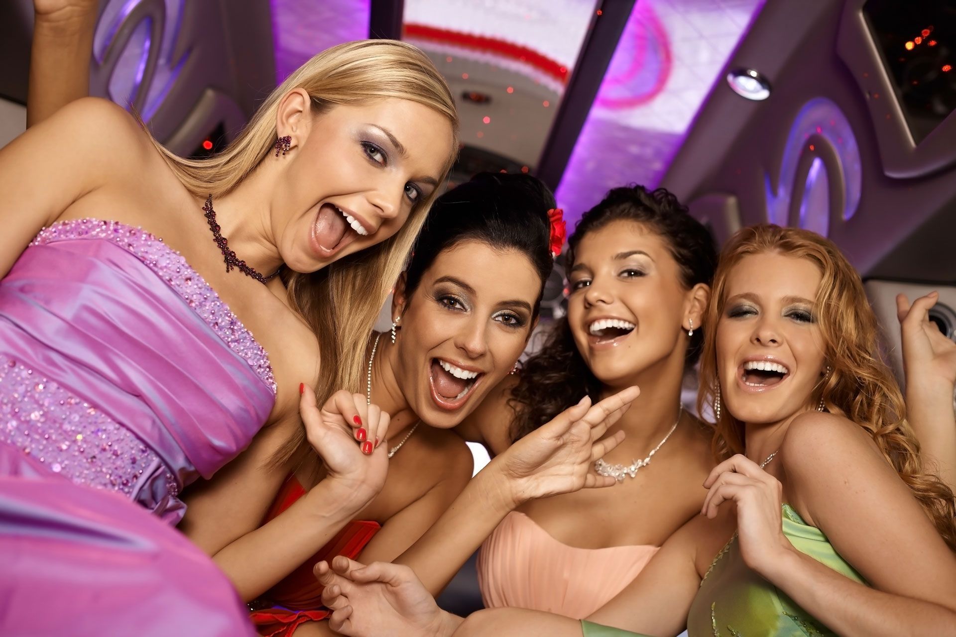 Four laughing people in formal dresses celebrate together inside a brightly lit limousine with purple ambient lighting.