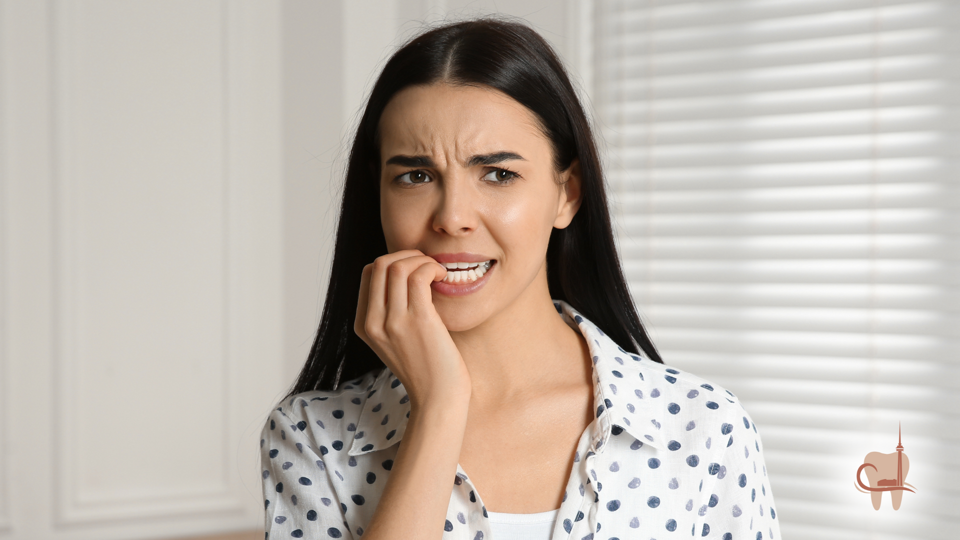 Woman with dark hair biting nails, looking anxious indoors, white dotted shirt.