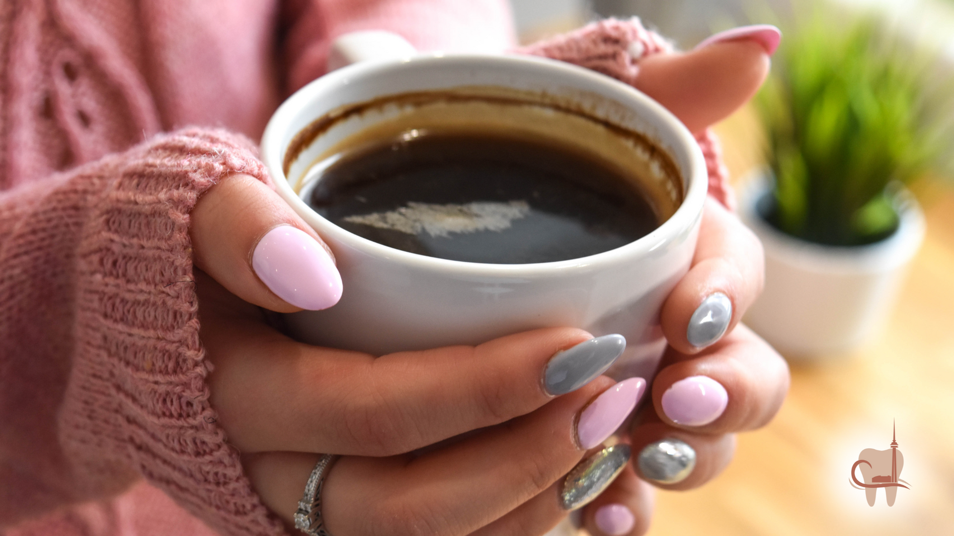 Woman's hands holding a cup of coffee, wearing pink and grey nail polish, pink sweater, indoors.