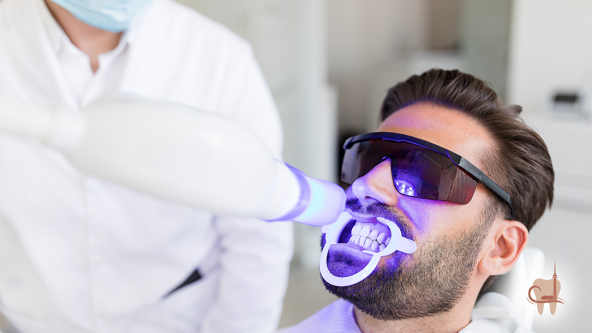 Man undergoing teeth whitening procedure at dentist's office, with UV light and protective eyewear.
