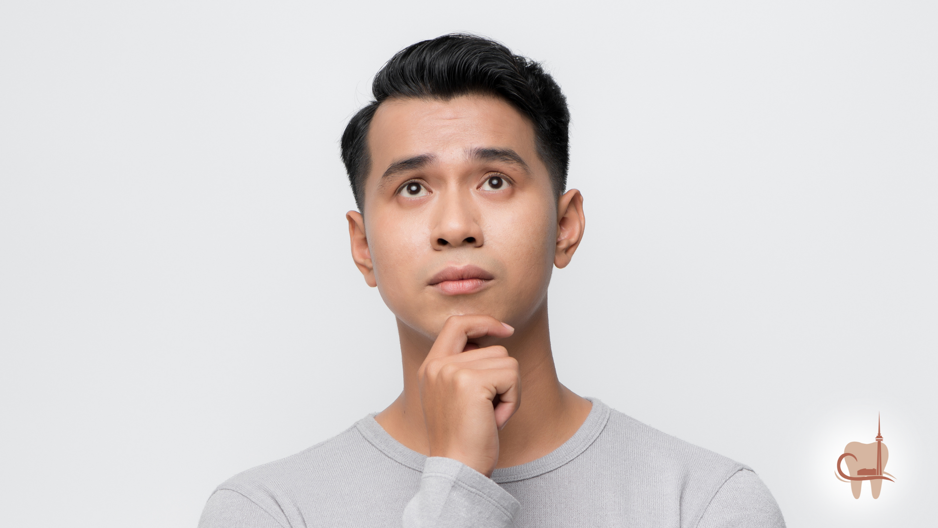 Man with dark hair looking upward, hand on chin, thoughtful expression, gray shirt, white background.