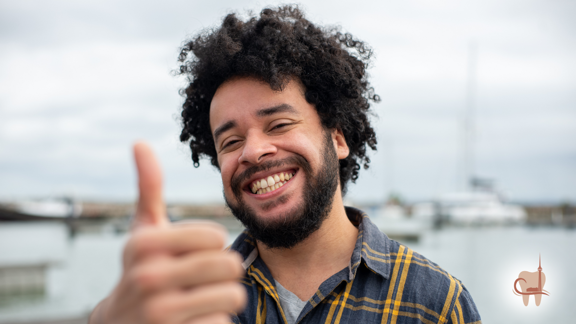 Man with curly hair smiling, giving a thumbs up in front of a harbor, cloudy sky.