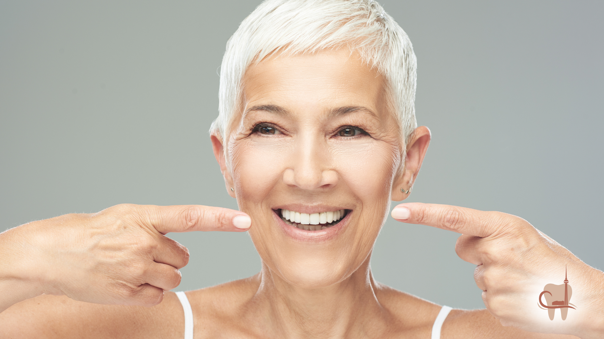 Smiling older woman with short white hair points to her teeth.