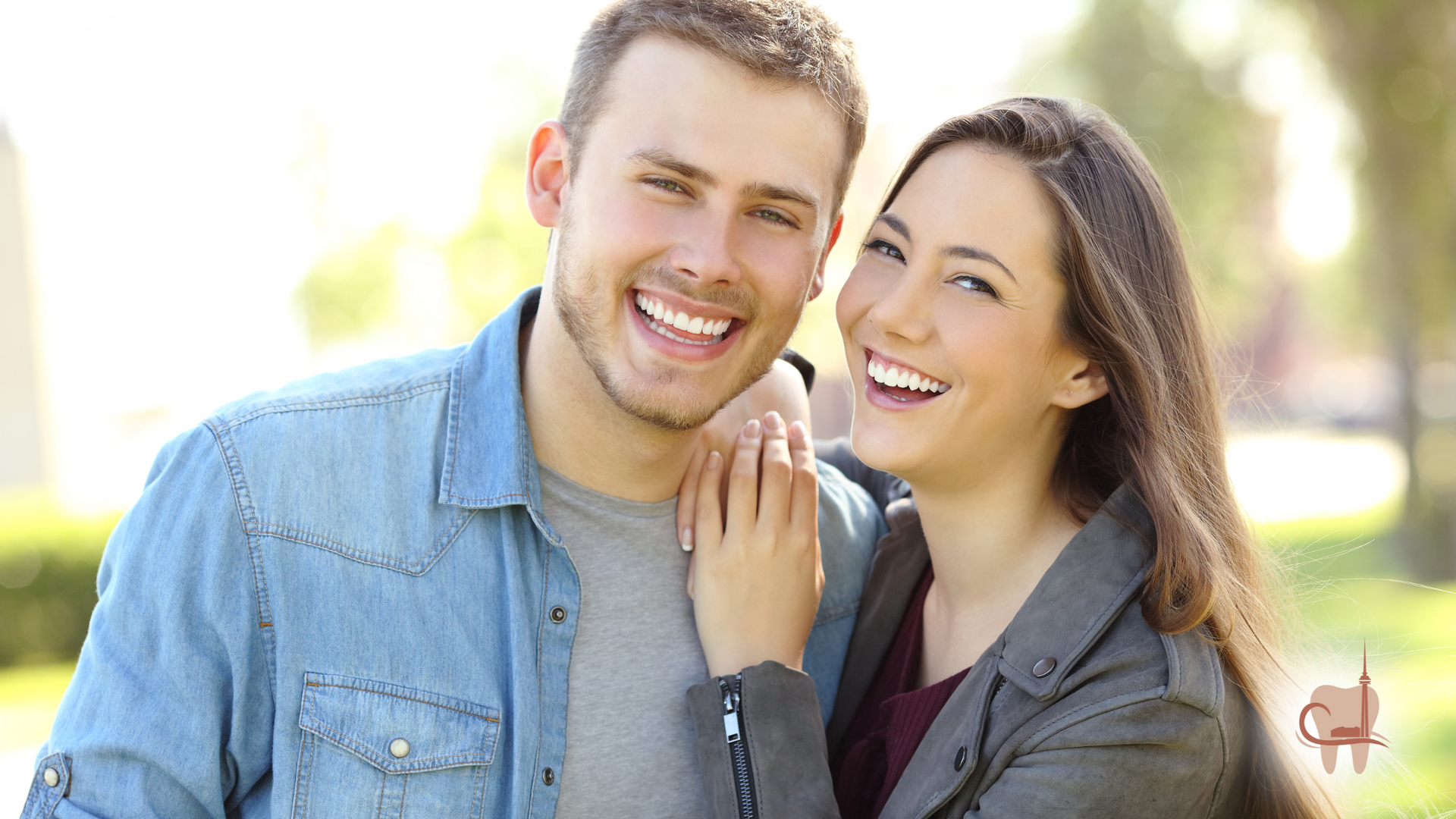 Smiling couple outdoors, man in denim shirt, woman with arm around him, sunny day.