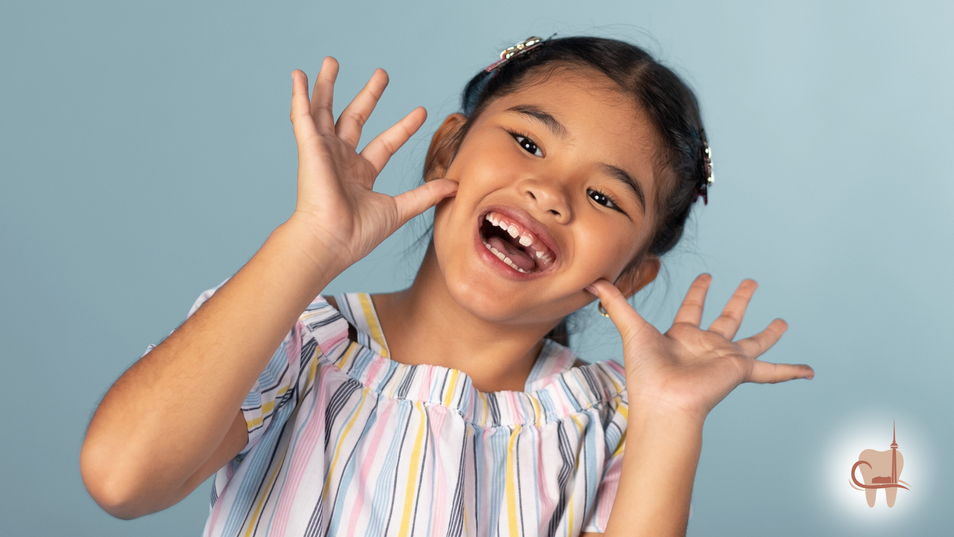 Girl with dark hair smiles, hands raised near her face, against a blue backdrop.