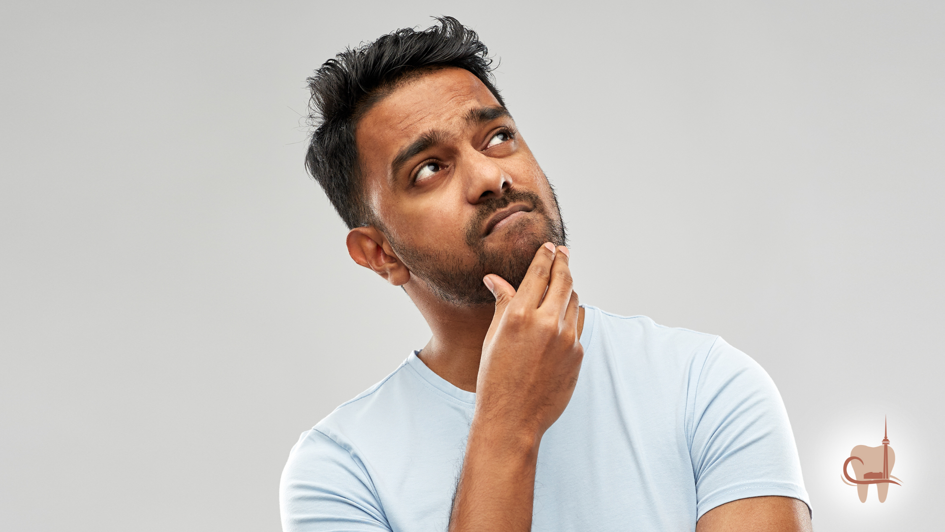 Man with a beard looking upward, hand on chin, pondering. Wearing a light blue t-shirt against a gray backdrop.